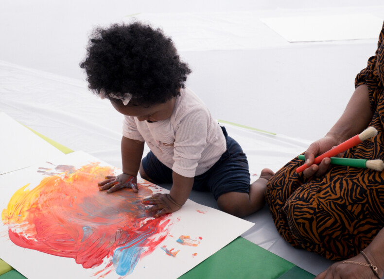 A toddler sat on the floor, painting with fingerpaint, hands full of blue, red and yellow paint.