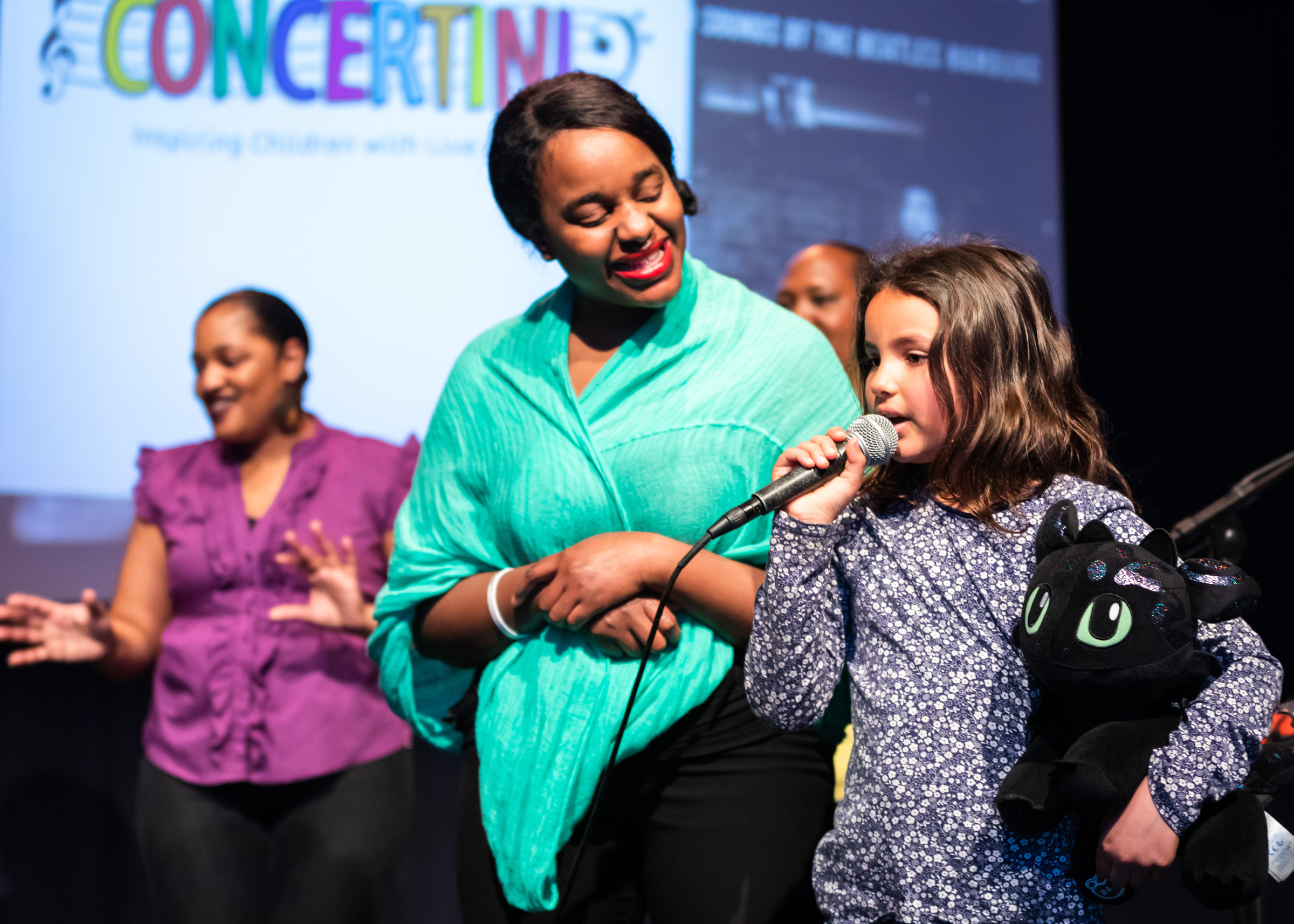 A child is speaking into a microphone at a concertini performance. An adult is standing beside the child, looking encouraging.