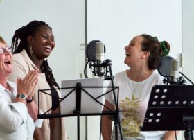 Three singers gathered around studio mics, laughing together