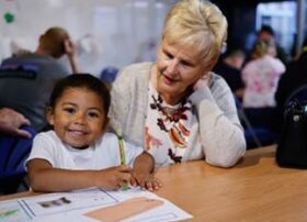 A child is colouring in an illustration. An elderly person is sitting nearby.