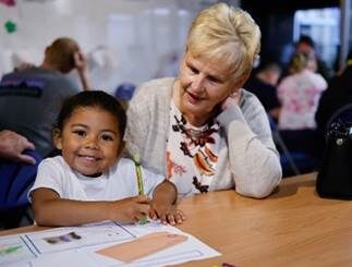 A child is colouring in an illustration. An elderly person is sitting nearby.