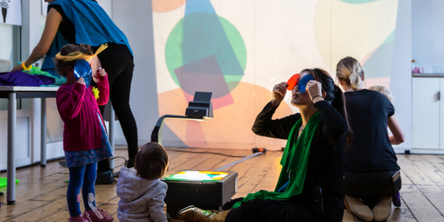 Person and baby hold coloured plastic over their eyes and project shapes onto the wall