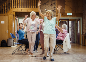 Elderly people dancing, both standing up and on chairs, representing age-friendly arts offers.
